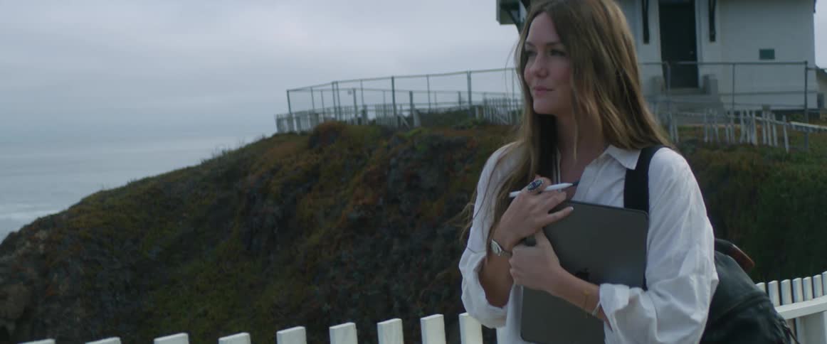 Woman walks on shore paralleling white picket fence with misty ocean and buildings in background