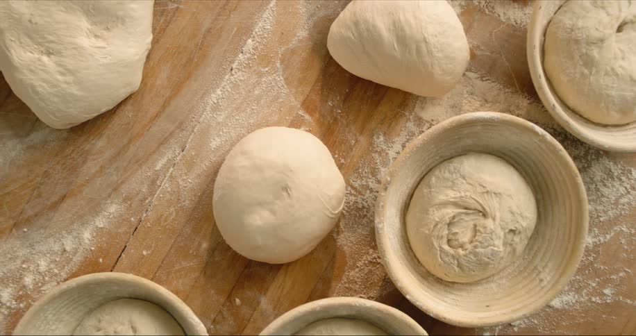 Several balls of dough sit on flour dusted wooden surface and in clay bowls