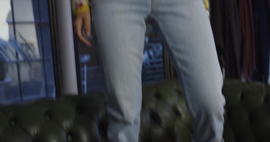 A girl is standing on a couch wearing white shoes and jeans with a yellow print blouse