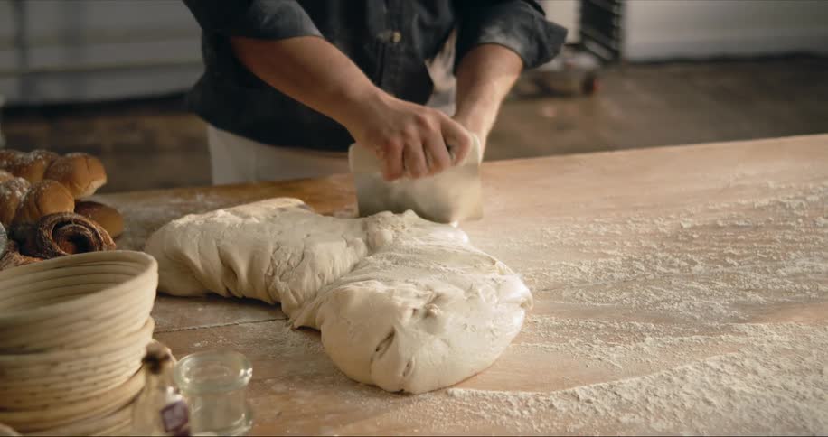 A man uses a pastry knife to cut dough on a large table