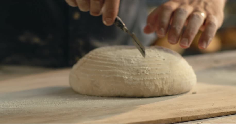 Baker man with black shirt scores the bread dough using a bread lame