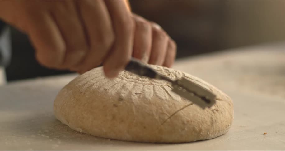 A man puts a decorative design on top of a loaf of bread and scores it before baking