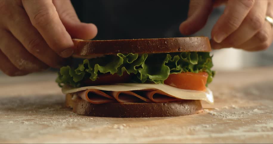 Man places top slice of bread on sandwich sitting on wooden surface dusted with flour