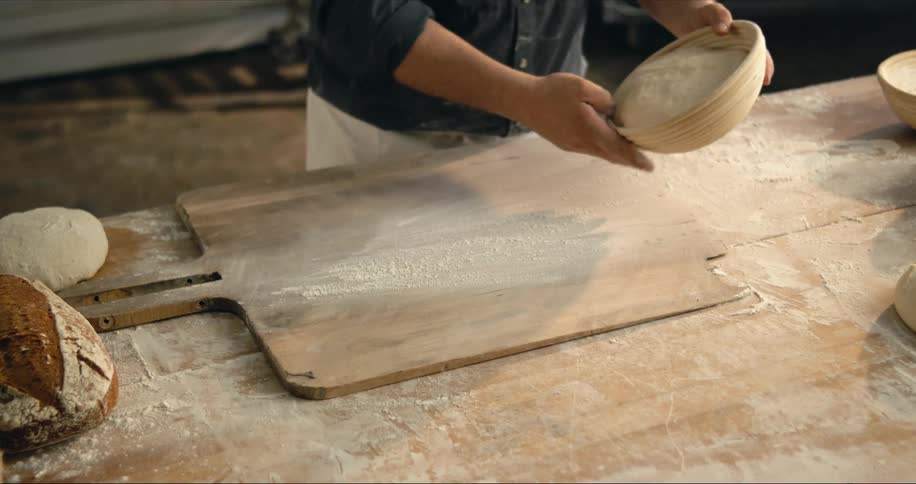 Man with apron around his waist dusts board with flour before placing dough from bowl onto it