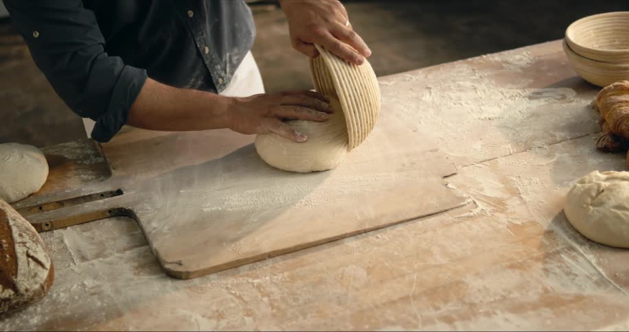 Baker pulls dough from bowl onto wooden board on flour dusted wooden table