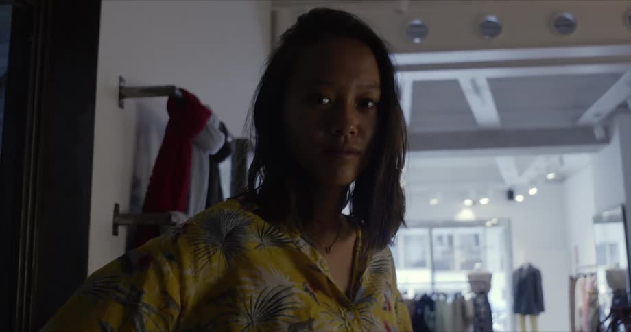 Woman with jeans yellow blouse and wavy hair stares down at camera in clothing shop