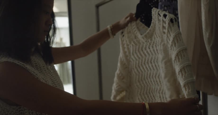 A woman in a clothing store taking a sweater from a rack with a mannequin a wall and window in the background
