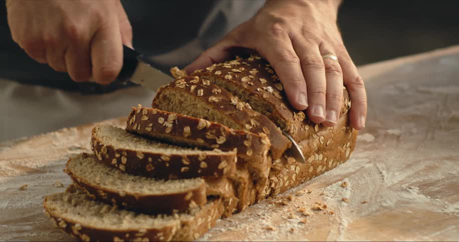 Man slices wheat bread sitting on flour dusted wooden surface using a bread knife