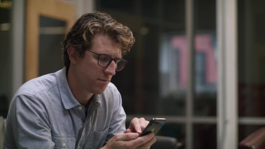 A man sits at a small table with a laptop and picks up his cell phone and looks at the screen
