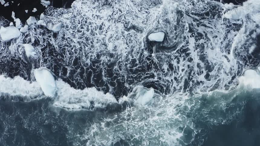 Aerial view of ocean waves crashing on the ice chunks over the beach