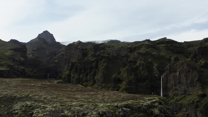 Aerial view of man standing on the edge of high cliff looking at high mountains with waterfall cascading down the mountainside