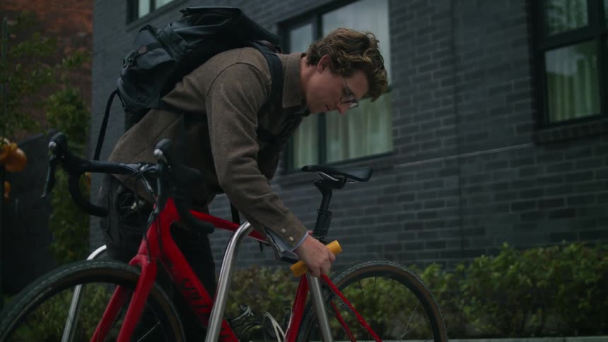 Man with glasses brown shirt and black backpack unlocks bike lock in front of apartment