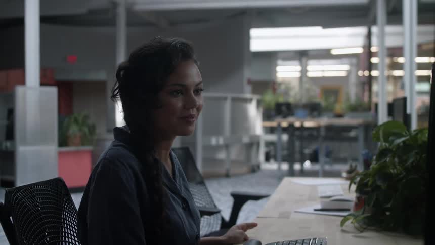 A woman in a room sitting on a chair at a desk in front of a computer with a wall in the background
