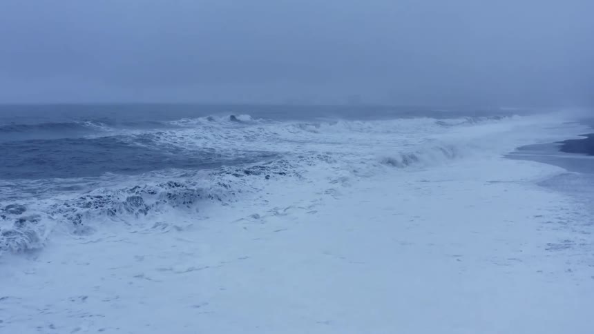 Waves crash onto beach creating foam under an overcast sky on foggy day