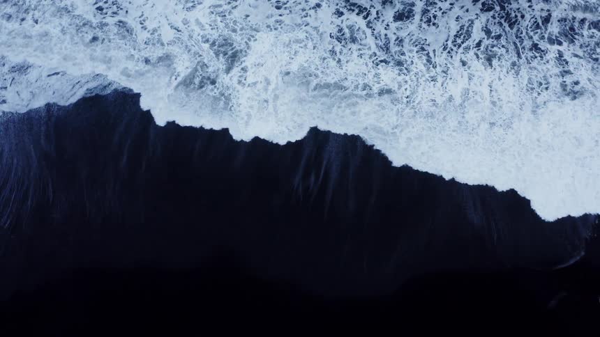 Aerial top down view of ocean waves crashing on the dark sand in the coast