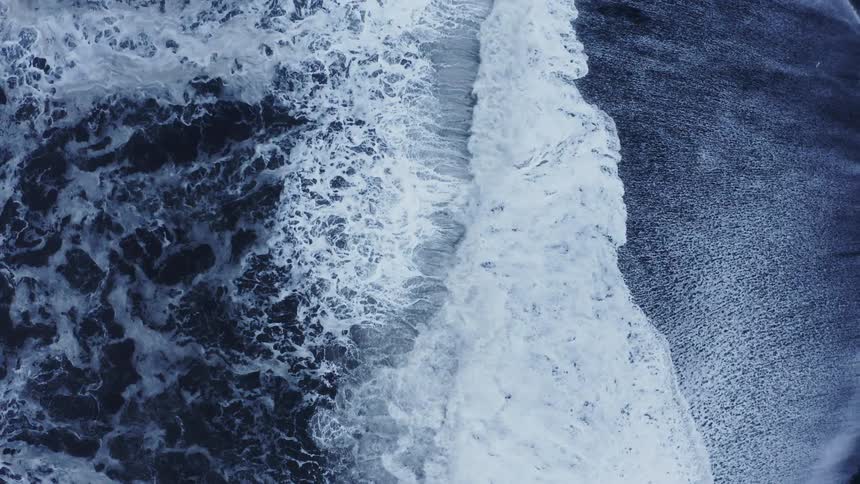 Aerial view of foamy ocean waves breaking against black sand beach