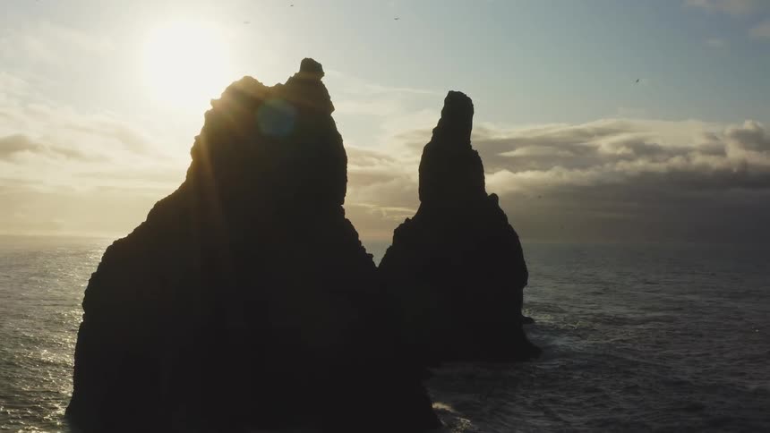 Silhouettes of rock formations in the water as the sun shines in a cloudy sky