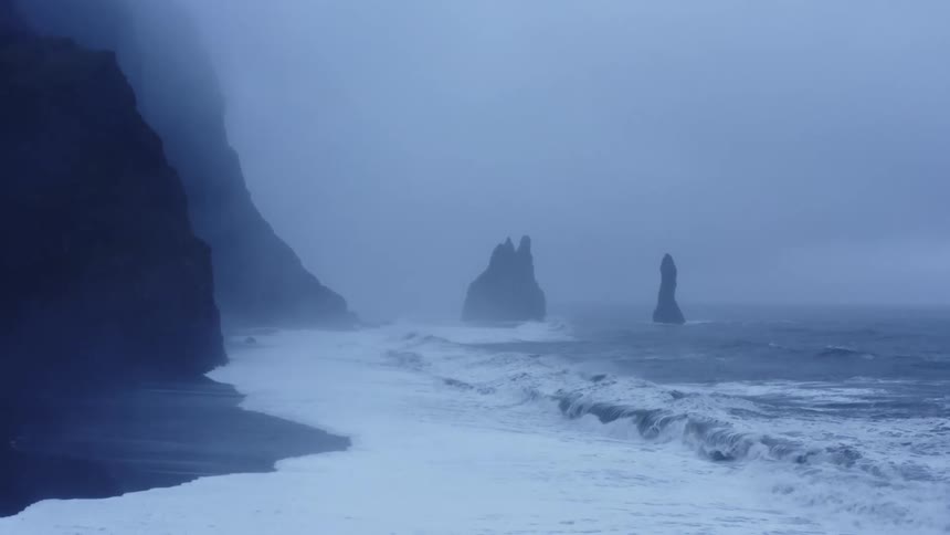 Ocean waves crash against the shore with cliff rocks on a foggy cloudy day