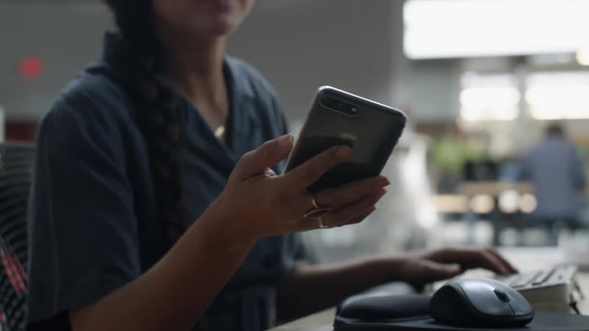 A woman sits at a desk typing on a computer keyboard and picks up her cell phone and smiles