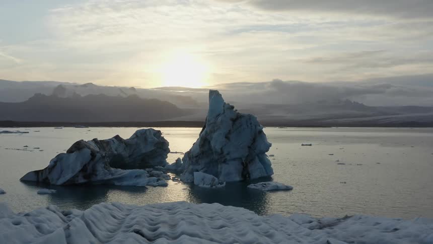 An aerial view of a large lake with floating icebergs with snowy mountains in the background in the early morning