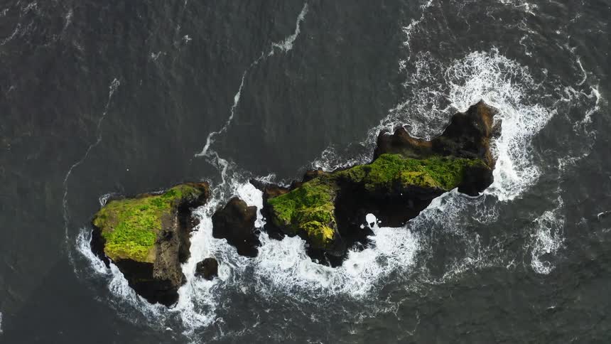 Birds fly around vegetation covered rocky island as waves crash against shore creating foam