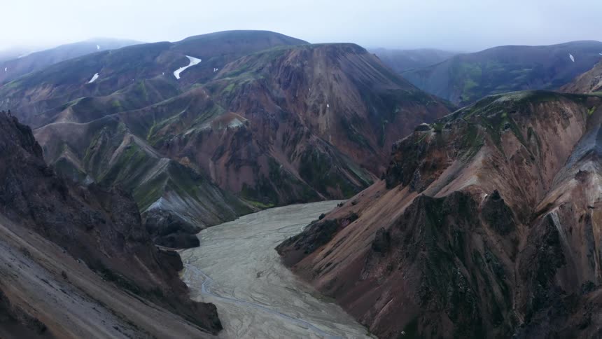 Small stream flows along wider riverbed winding through vegetation dotted mountainous terrain as fog covers summits and plateaus on horizon