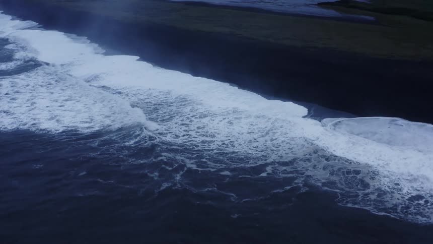 Aerial view of foamy ocean waves breaking against black sand beach