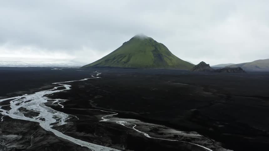 Pale reflective streams flow toward vegetation covered mountain with fog on horizon under overcast sky