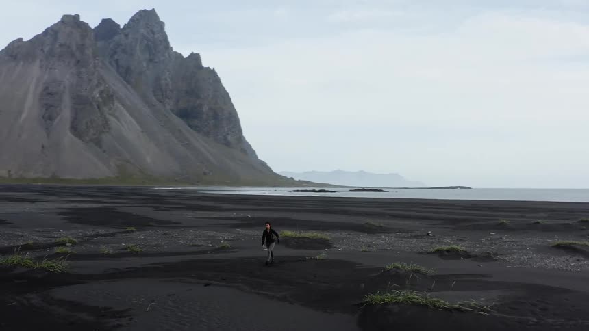 Woman stands on black sand beach looking out to sea before turning and jogging away with craggy mountain in background