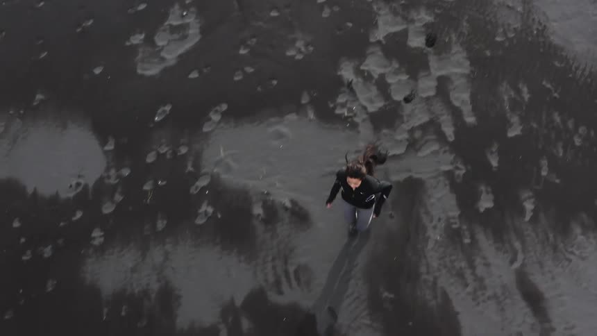 Aerial view of woman with ponytail black jacket and gray leggings running on black sand beach