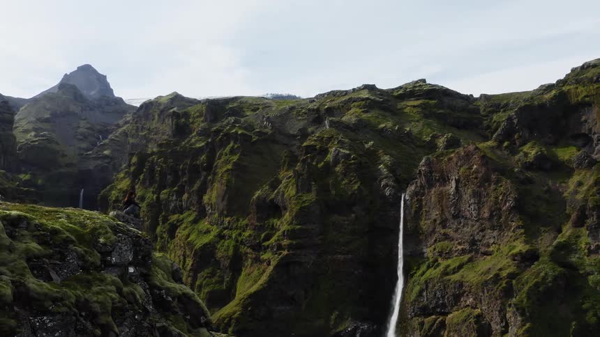 A person sits on a cliff rock ledge looking out over the ravine at a waterfall