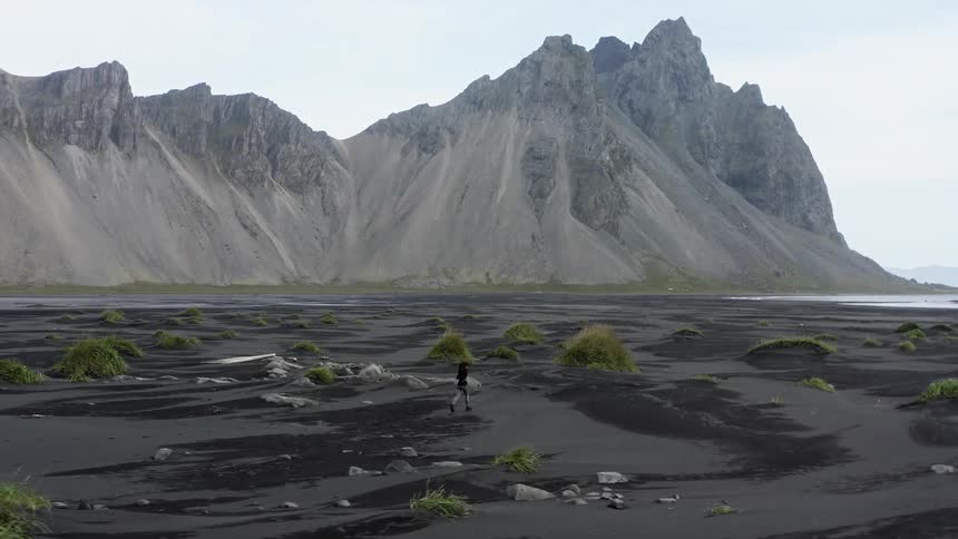 A woman is running across land towards water with mountains nearby
