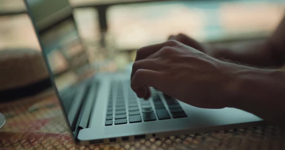 A man sits on a balcony and types on his laptop and then answers his cell phone and talks