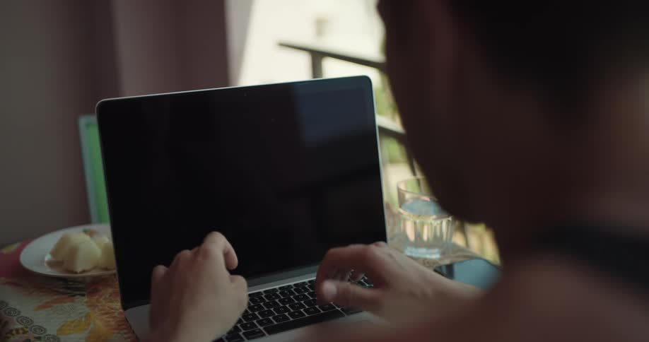A man is sitting at a small table in an apartment kitchen while typing on a laptop near a window during the day