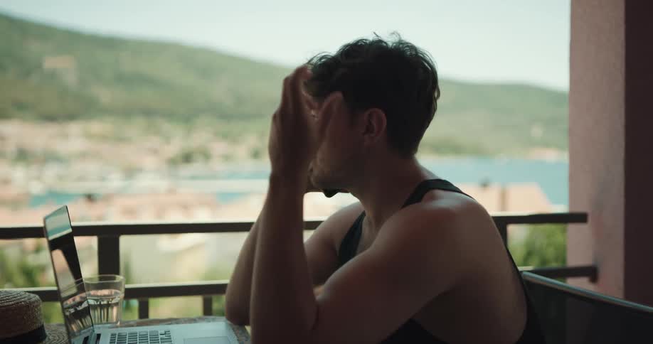 A man is sitting at a table on a balcony with a laptop computer in front of him as he talks on his phone