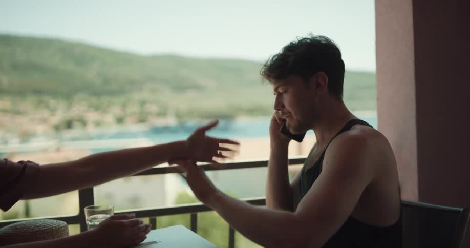 A man and a woman on a balcony arguing with the man talking on a smartphone with the sky and clouds in the background
