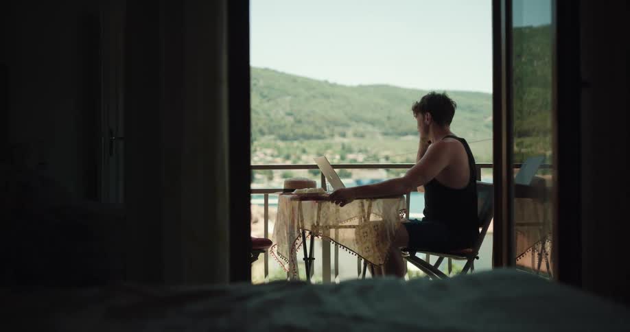 A man sitting on a chair next to a table and a laptop on a balcony with the sky and clouds in the background