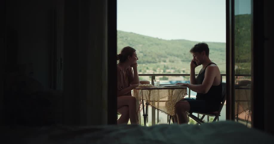 Couple share food sitting on table on balcony with view of hill and water