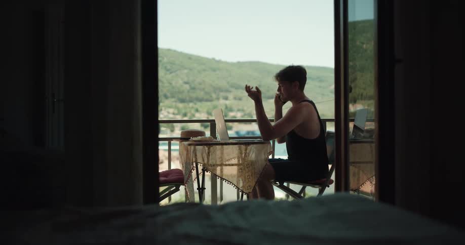 Young man sitting at a table on the apartment balcony talking on the phone