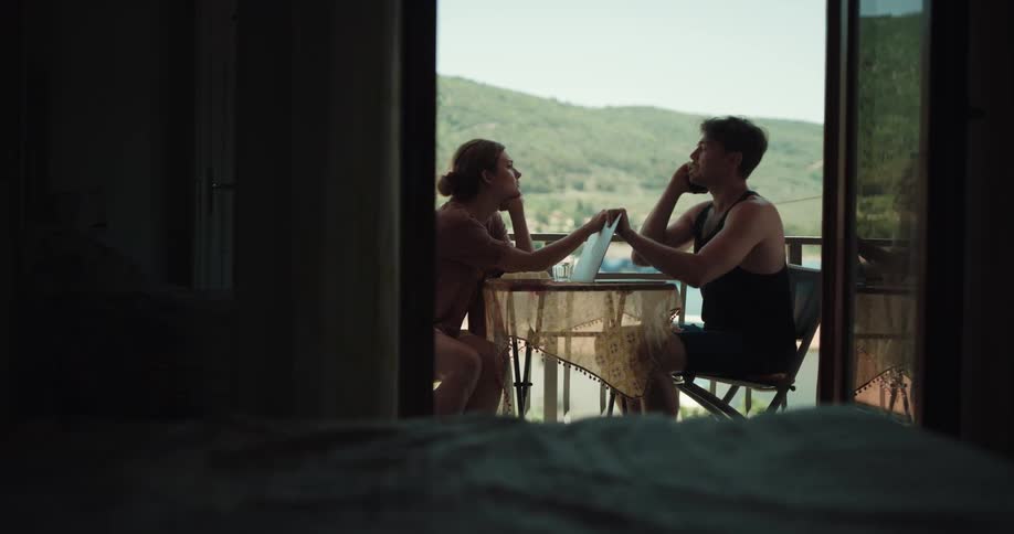 A man sitting on a chair at a table on a balcony with a woman teasing him with hills the sky and clouds in the background