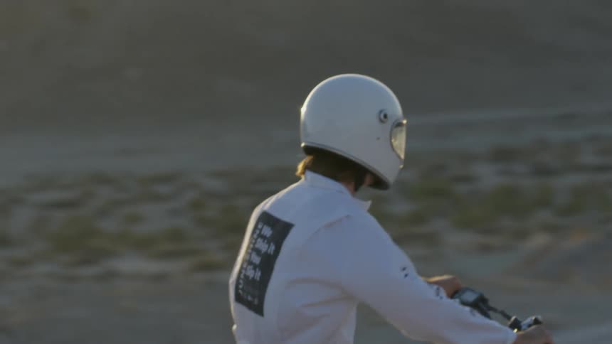 A man wearing white coveralls and a helmet rides a motorbike in the desert with sunlight reflecting off his visor