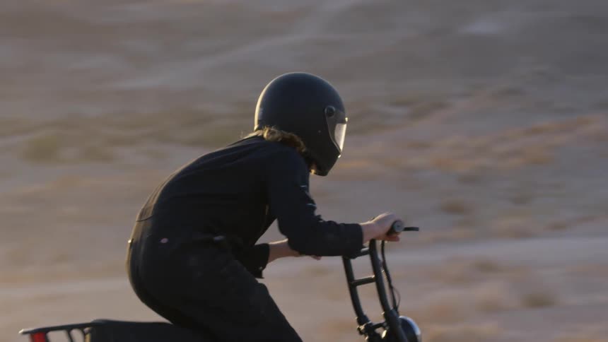 A cyclist dressed in black and wearing a black helmet rides a motorbike in the desert