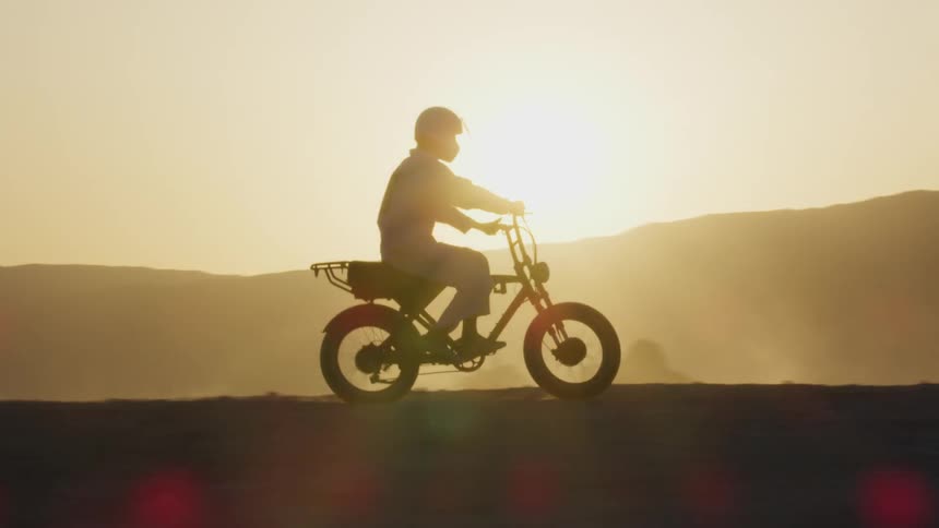 Silhouette of cyclist riding a motorbike with a bright sun over mountains on the horizon