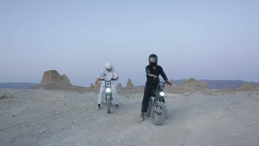 Two people on motorcycles in a desert with the sky and clouds in the background