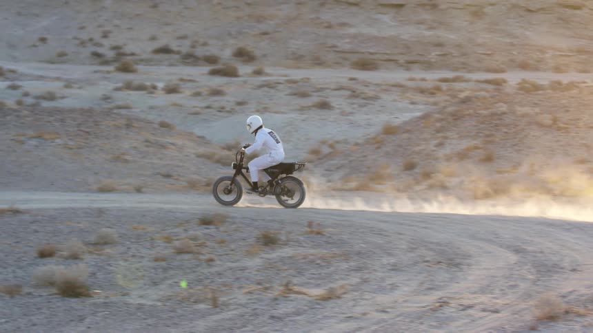 A cyclist dressed in white and wearing a white helmet rides a motor bike in the desert
