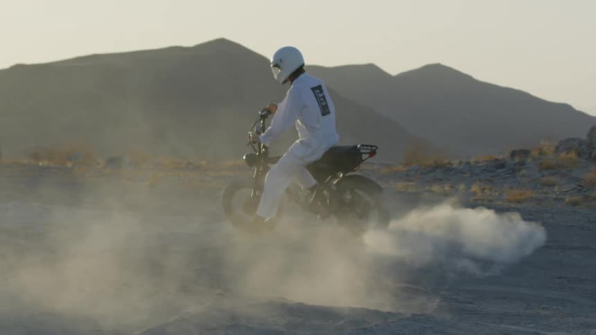 A cyclist dressed in white and wearing a white helmet rides a motorbike in the desert with mountains in the distance