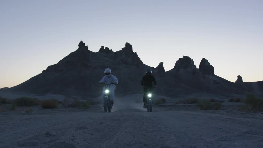 Two motorcycles travel across a desert with a mountains behind them