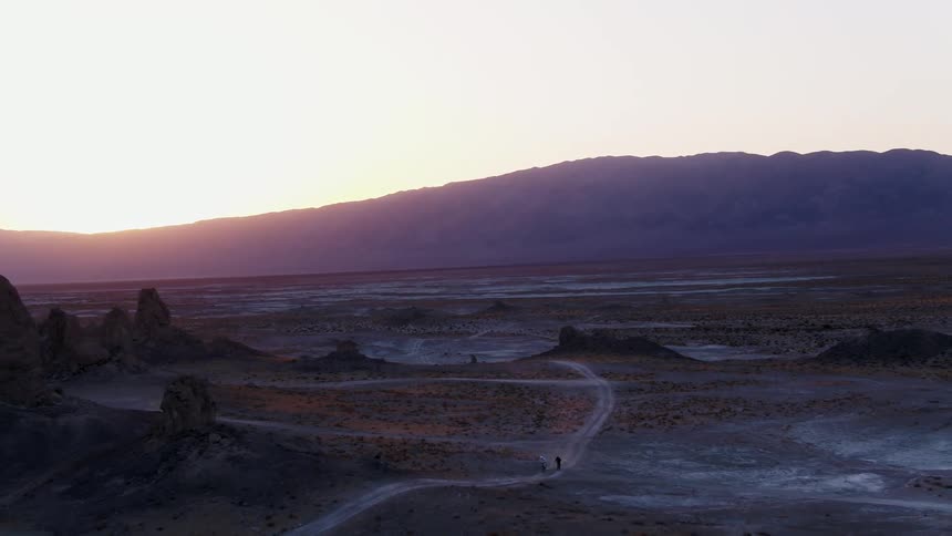 Two people ride motorbikes on a narrow dirt trail through barren land at sunset