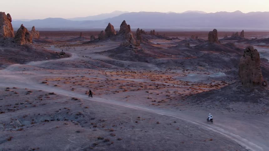 Two people riding motorcycles on a dirt road in a desert landscape with the sky and clouds in the background