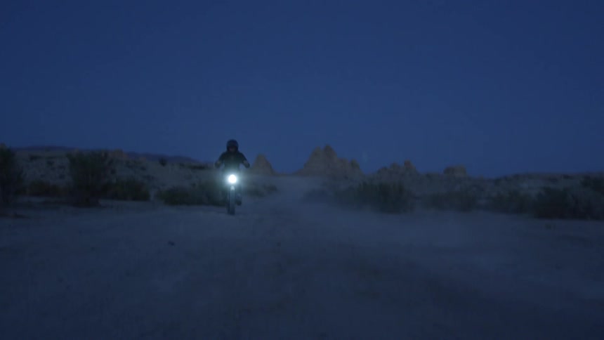 Two people are riding electronic bikes on a sand path in the desert after dusk